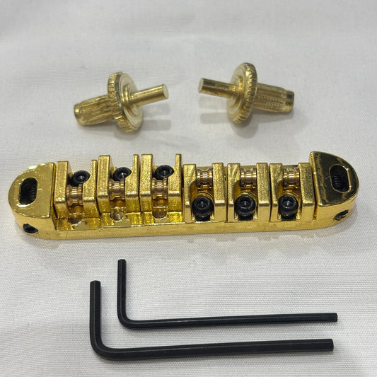 Gold electric guitar roller bridge with two supports pins and a hex key on a white background.