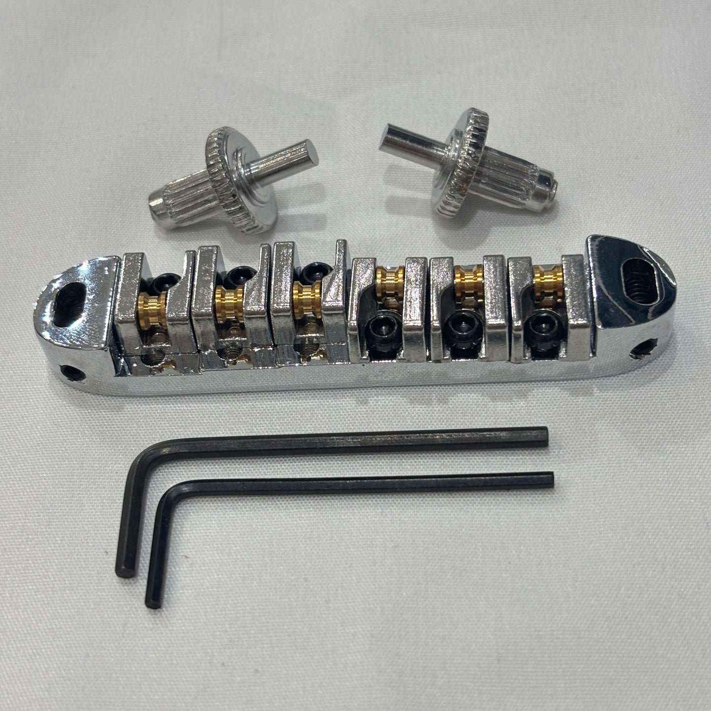 Chrome electric guitar roller bridge with two supports pins and a hex key on a white background.