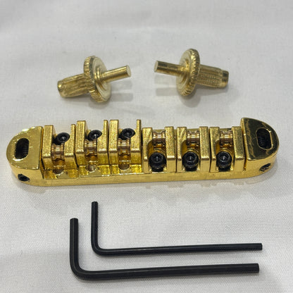 Gold electric guitar roller bridge with two supports pins and a hex key on a white background.