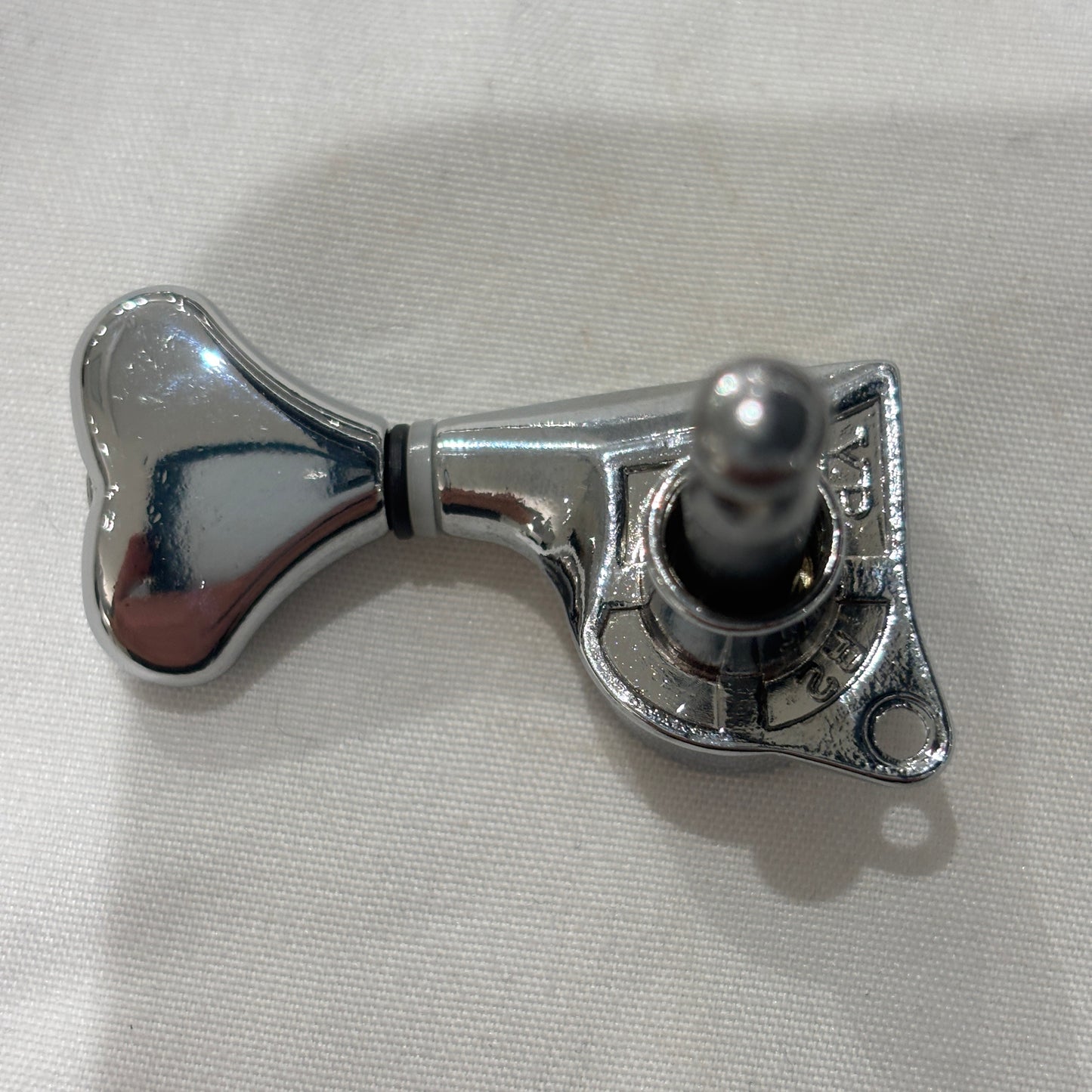 The back of a Chrome Fish Tail Button Guitar Tuner on  textured fabric background.