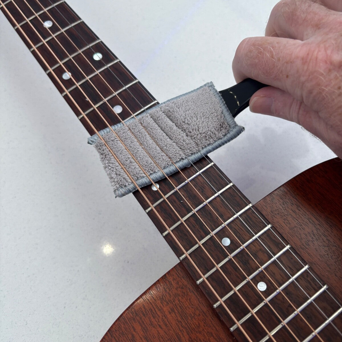 Person cleaning a guitar neck with a microfibre pad on a grey surface.
