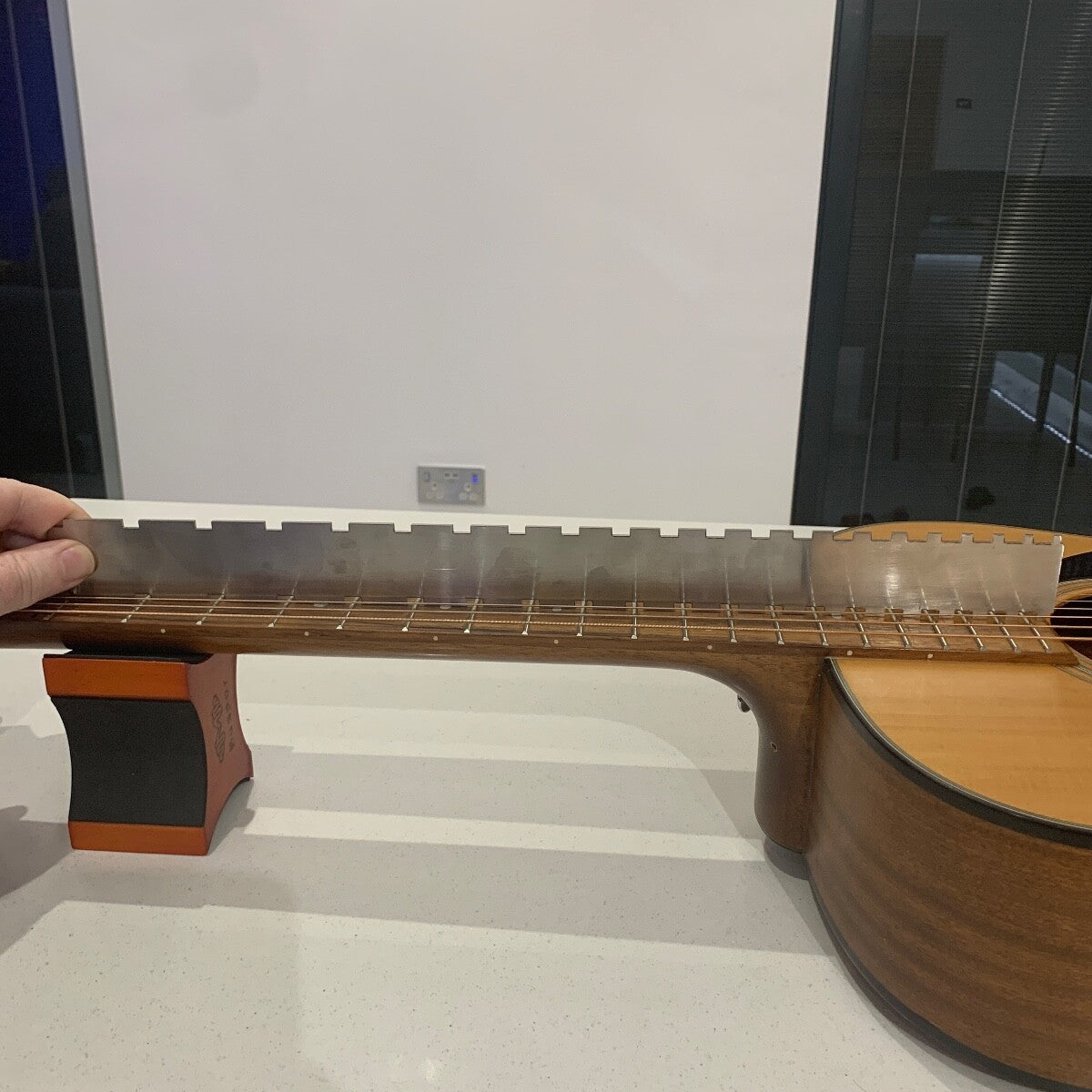Close-up of a Guitar Notched Straight Edge being used on the neck of a guitar with a hand holding it on a light-coloured surface.